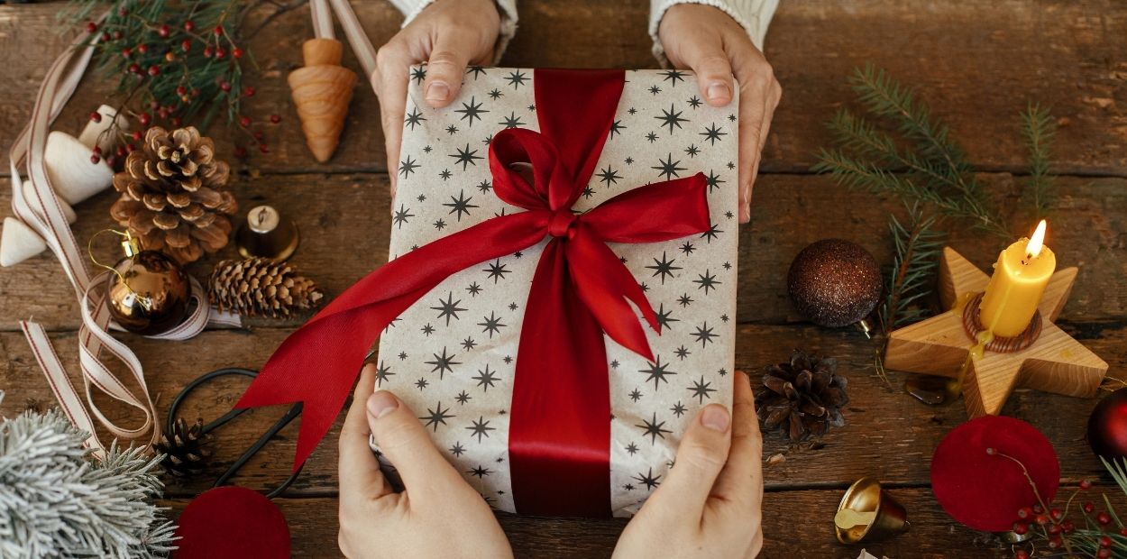 A giftbox wrapped in white paper with silver stars and a red box is being passed from one set of hands to another. On the wooden table under the gift there is a collection of festive items including pinecones, candles, bells, ribbon, and greenery.